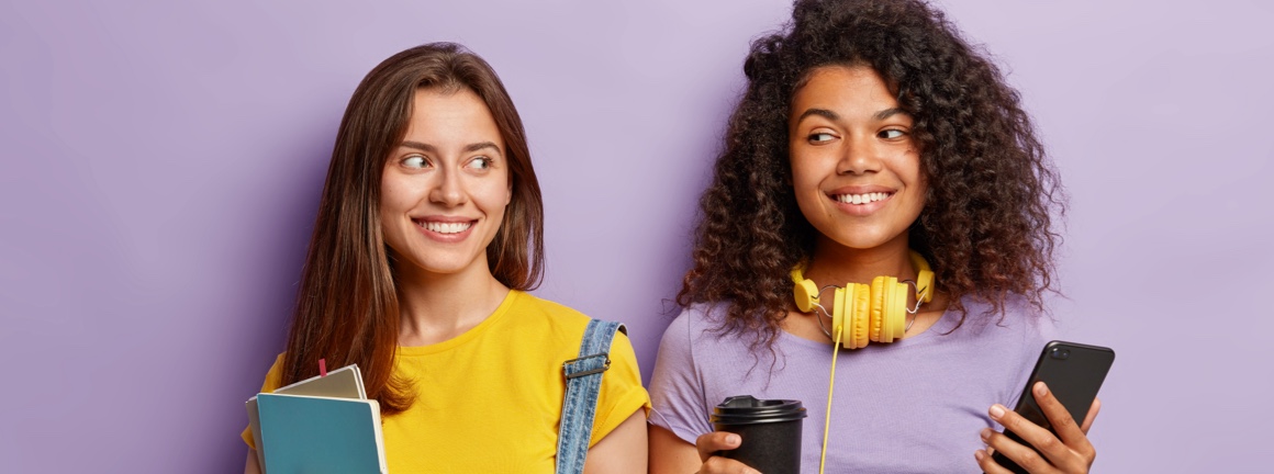 Two teenage girls smiling while standing beside each other with books, coffee and phones in hand.